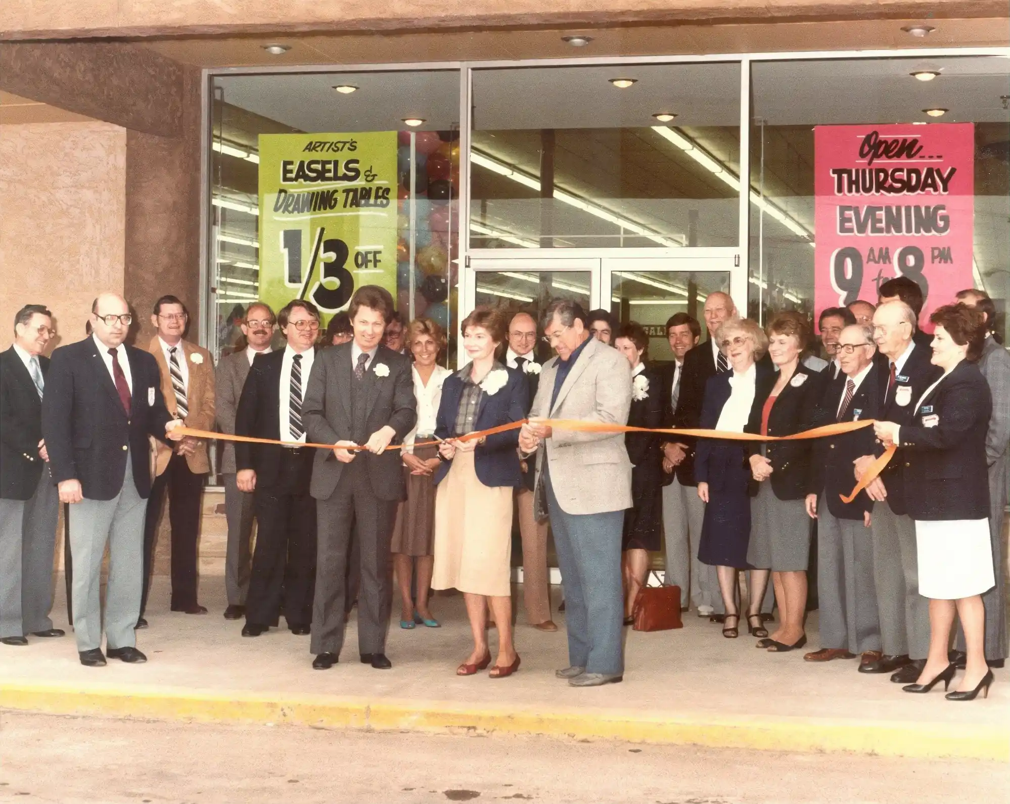 The opening ceremony of the first Hobby Lobby store. David and Barbara Green stand in the center of a crowd of people. The Green couple are holding scissors to cut the ceremonial ribbon draped in front of them.